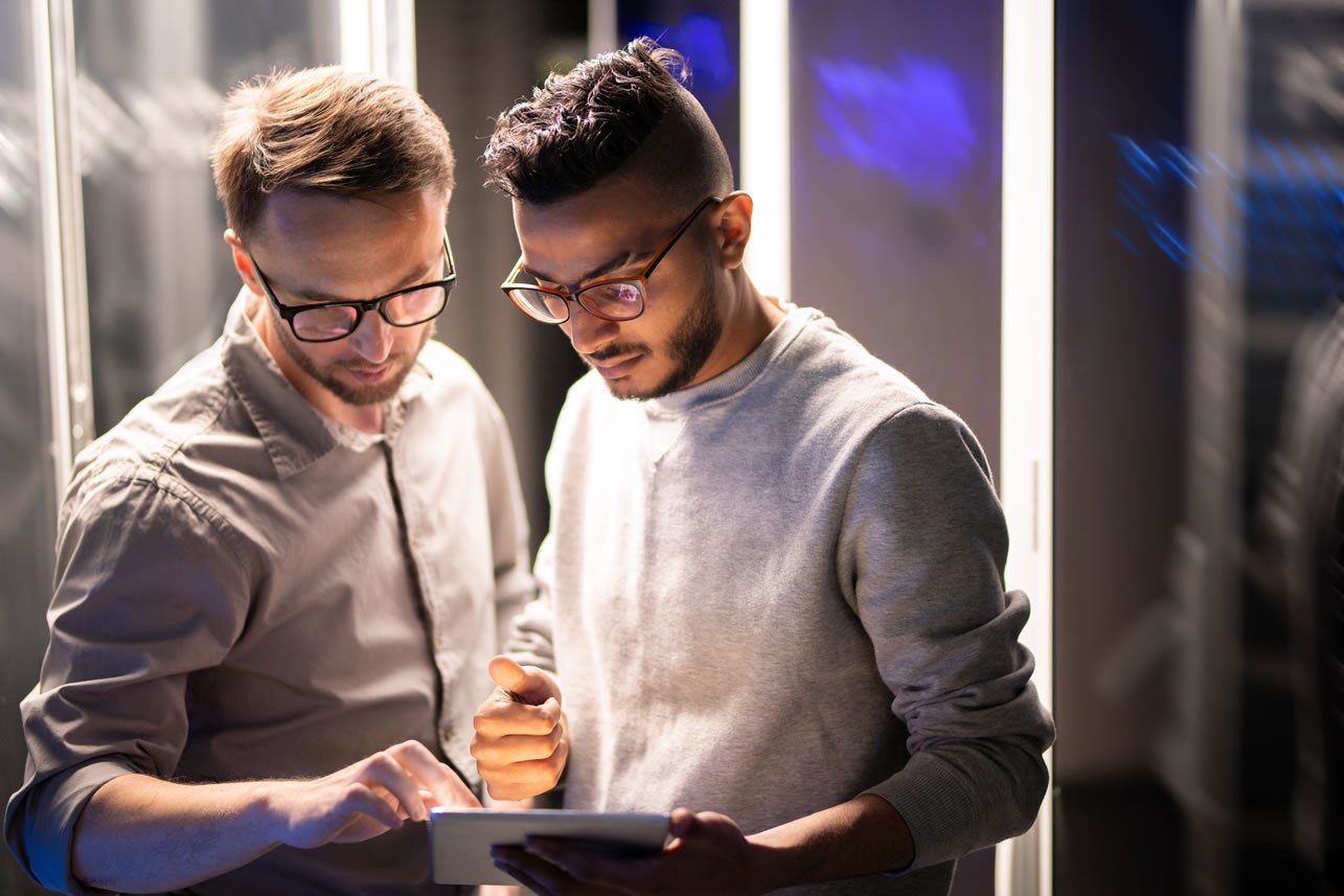 Two network security specialists conferring over a tablet in a server room